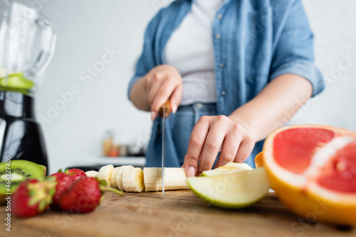 Fototapeta Naklejka Na Ścianę i Meble -  Cropped view of blurred woman with overweight cutting banana near fruits and blender in kitchen