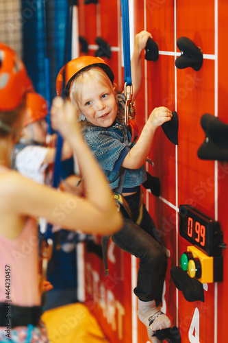 Indoor climbing activities for kids. School b oy smiling at the camera and having fun in indoor playground for children. Happy kids in red helmets climbing the wall in bouldering center