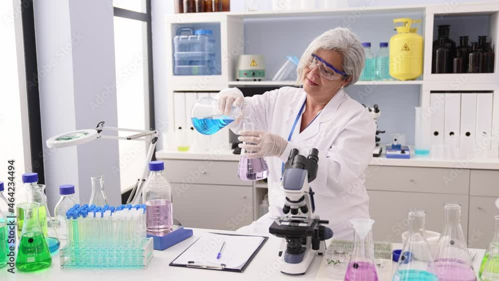 Middle age grey-haired woman wearing scientist uniform mixing liquid working at laboratory Middle age grey-haired woman wearing scientist uniform mixing liquid working at laboratory