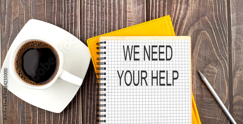 WE NEED YOUR HELP text on the notebook with coffee on wooden background