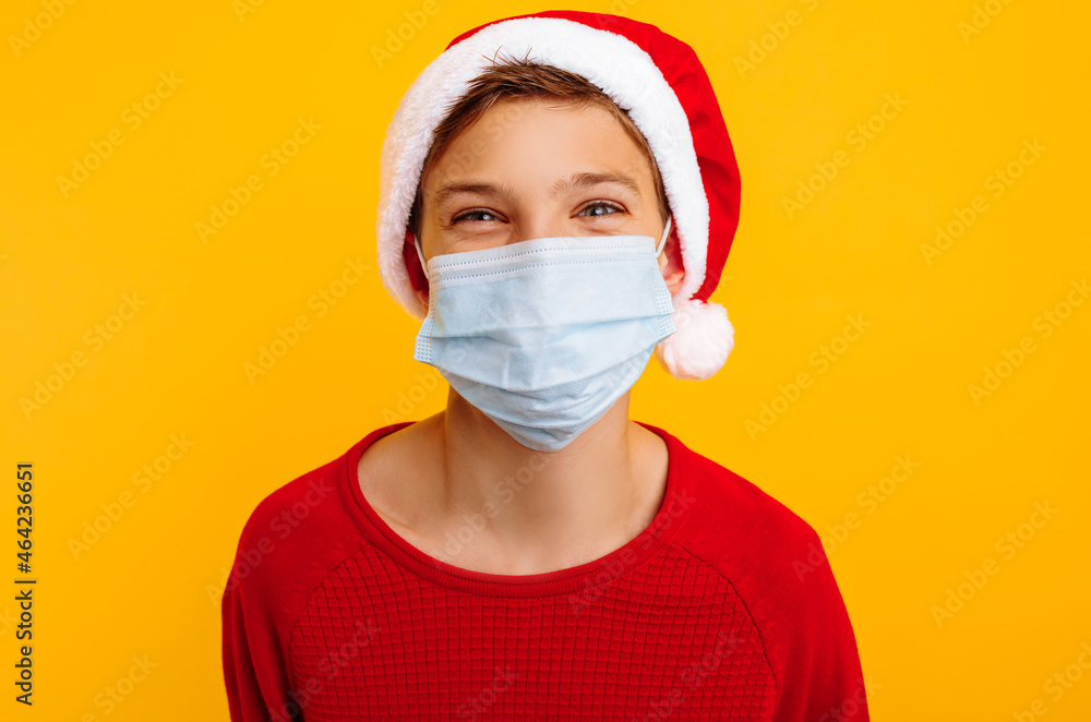 Happy boy in a protective mask and santa claus hat, on a yellow background. Protection from virus. pandemic COVID 19 Happy boy in a protective mask and santa claus hat, on a yellow background. Protection from virus. pandemic COVID 19