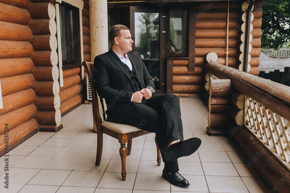 A stylish, young, rich male businessman in a black suit sits on a wooden chair on the veranda of his house. Portrait of a fashionable successful man. A stylish, young, rich male businessman in a black suit sits on a wooden chair on the veranda of his house. Portrait of a fashionable successful man.