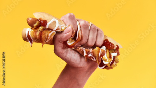 Female hand squeezing junk hot dog with mustard and ketchup isolated over yellow background