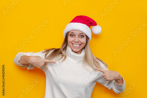 A young smiling blonde woman in  a white sweater and Christmas Santa hat points with fingers, showing empty copy space for design and text isolated on a color yellow background © Марина Демешко