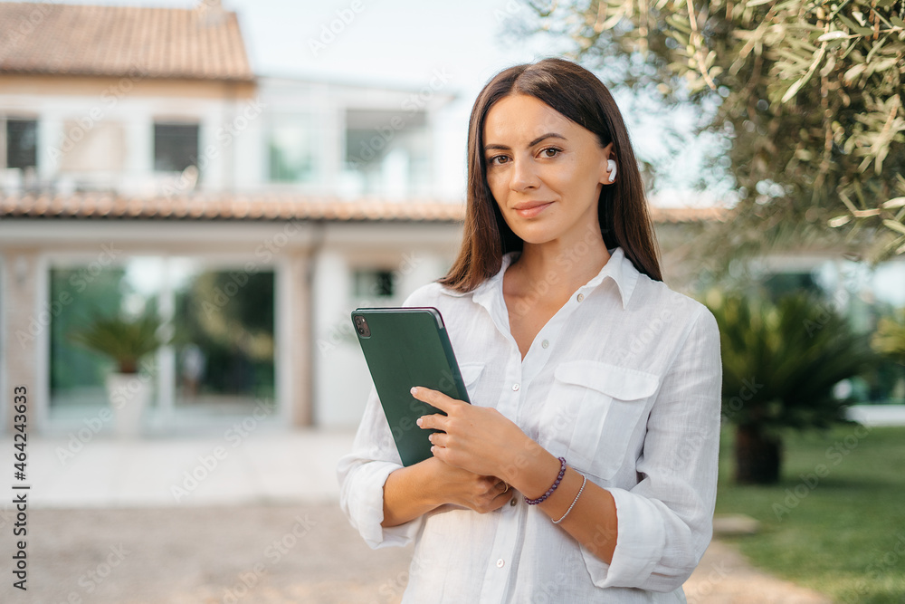 Portrait of a happy brunette woman smiling holding a tablet in the hands of a summer garden white shirt. The realtor presents and sells the house Portrait of a happy brunette woman smiling holding a tablet in the hands of a summer garden white shirt. The realtor presents and sells the house