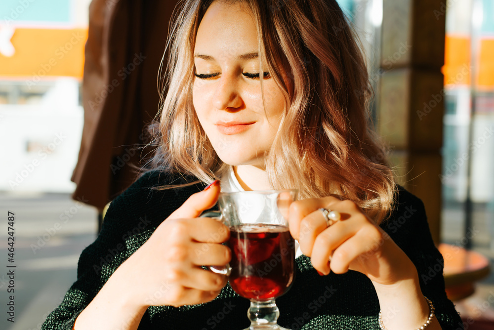 Portrait of young woman with glass of mulled wine and closed eyes enjoying rest, break in cafe or bar. Close-up of tenderly smiling girl's face inside room Portrait of young woman with glass of mulled wine and closed eyes enjoying rest, break in cafe or bar. Close-up of tenderly smiling girl's face inside room