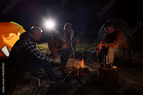 Group of friends with guitar near bonfire and camping tent outdoors at night