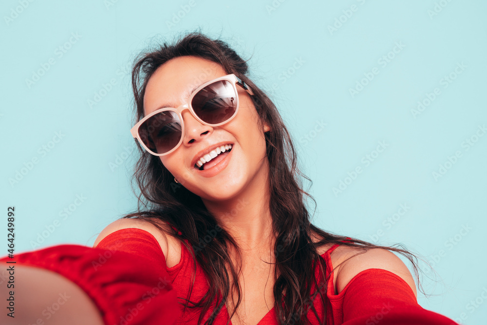 Fototapeta premium Young beautiful smiling female in trendy summer red top and jeans clothes. Sexy carefree woman posing near blue wall in studio. Positive brunette model having fun. Taking Pov selfie in sunglasses