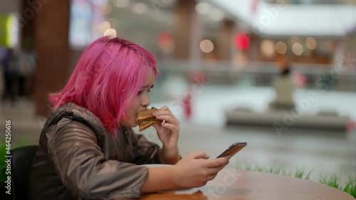 A focused schoolgirl with pink hair, holding a mobile phone in her hands, eats bread and sausage in a shopping center cafe photo