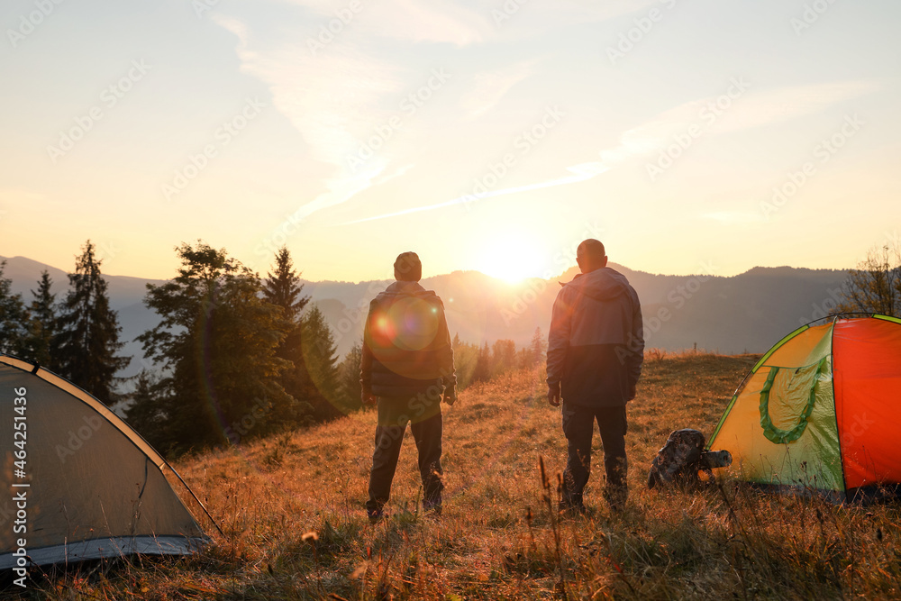 Men near camping tents in mountains at sunset, back view
