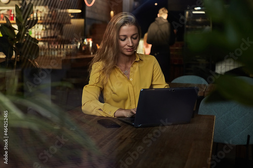 beautiful young caucasian business woman working on laptop. female freelancer in yellow shirt sitting at table, shot through window glass outside of cafe. modern technology, distant work, remote job
