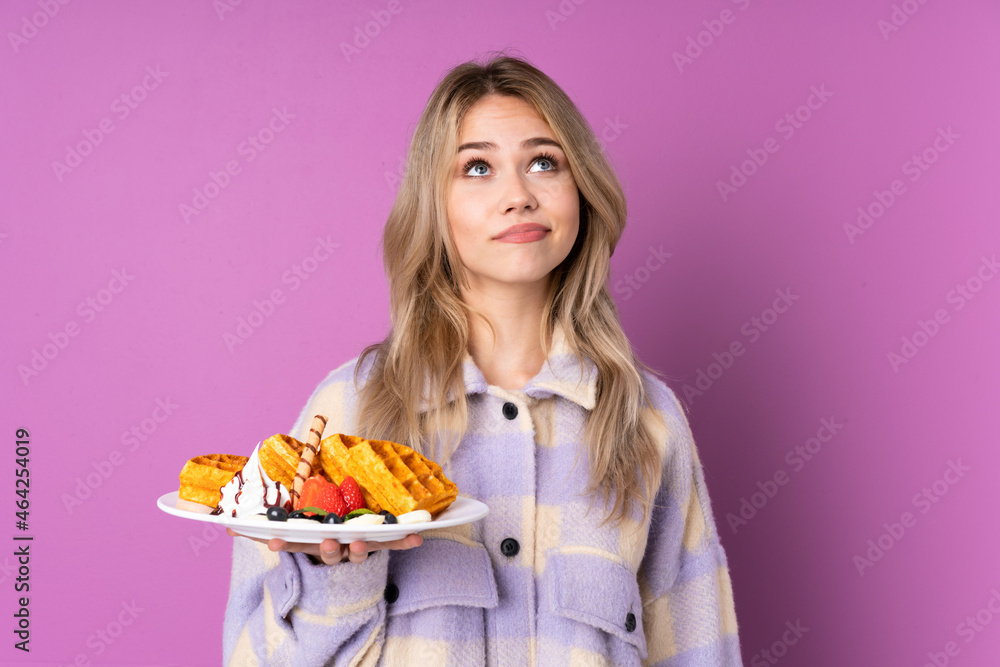 Teenager Russian girl holding waffles isolated on purple background and looking up