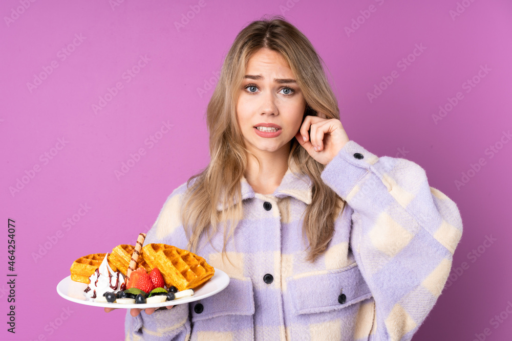 Teenager Russian girl holding waffles isolated on purple background frustrated and covering ears Teenager Russian girl holding waffles isolated on purple background frustrated and covering ears