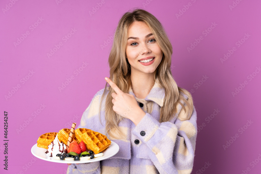 Teenager Russian girl holding waffles isolated on purple background pointing to the side to present a product Teenager Russian girl holding waffles isolated on purple background pointing to the side to present a product