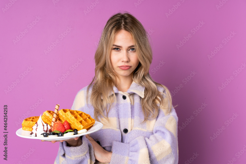 Teenager Russian girl holding waffles isolated on purple background feeling upset