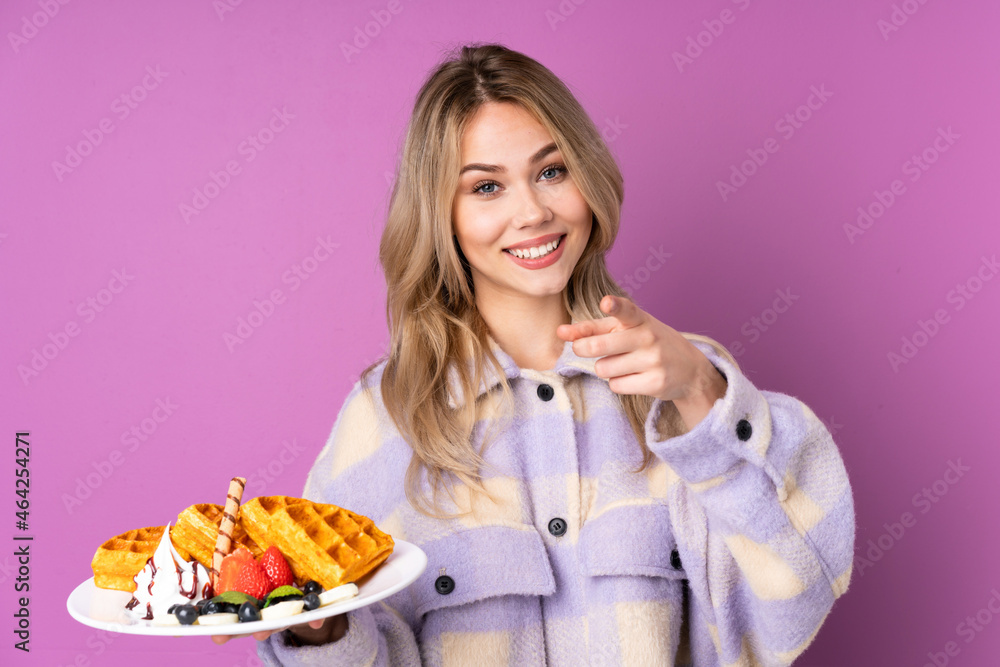 Teenager Russian girl holding waffles isolated on purple background pointing to the front and smiling Teenager Russian girl holding waffles isolated on purple background pointing to the front and smiling