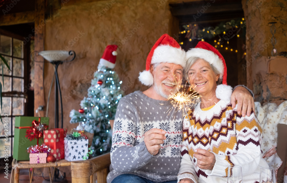 Defocused senior couple in Santa hats hugging at home at Christmas time sparkling. Sitting inside a rustic mountain chalet with Christmas tree, presents and lights on background Defocused senior couple in Santa hats hugging at home at Christmas time sparkling. Sitting inside a rustic mountain chalet with Christmas tree, presents and lights on background