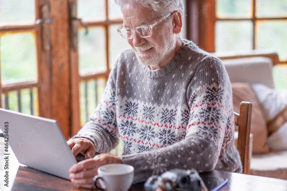 Smiling white haired senior man in winter sweater sitting in living room using laptop computer. Carefree elderly grandfather enjoying tech and social. Rustic chalet in the wood Smiling white haired senior man in winter sweater sitting in living room using laptop computer. Carefree elderly grandfather enjoying tech and social. Rustic chalet in the wood