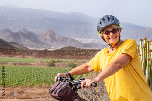 Old senior woman enjoying outdoor excursion with bicycle and healthy lifestyle. Elderly smiling woman wearing sport helmet in countryside with vineyard and mountain