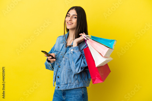 Young caucasian woman isolated on blue background holding shopping bags and writing a message with her cell phone to a friend