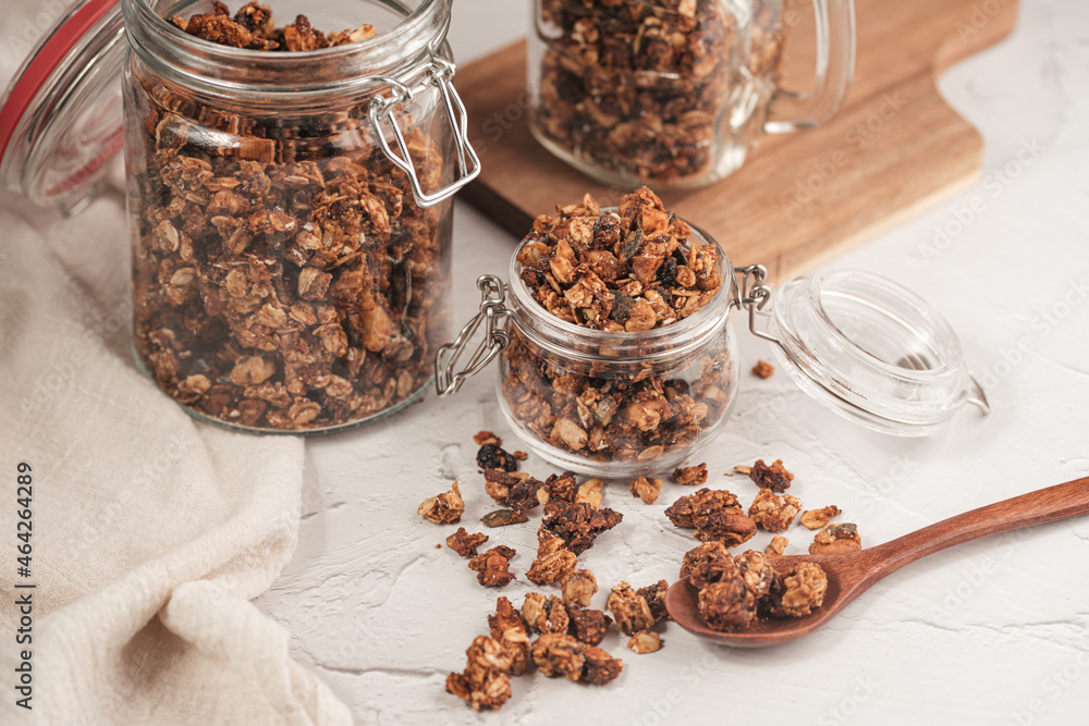 Mason jar with granola breakfast cereal with nuts. wooden spoon with scattered granola in front and two jars with granola on small wooden board in the back on the off-white surface Mason jar with granola breakfast cereal with nuts. wooden spoon with scattered granola in front and two jars with granola on small wooden board in the back on the off-white surface