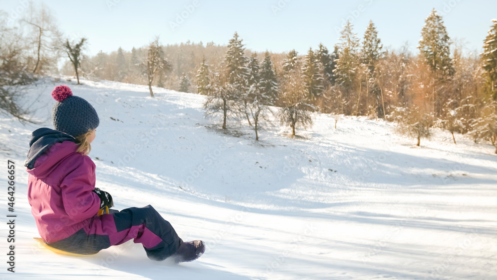 Llittle girl sledding with a warm knit hat, laughing descending the icy slope on the yellow shovel sledge. Llittle girl sledding with a warm knit hat, laughing descending the icy slope on the yellow shovel sledge.