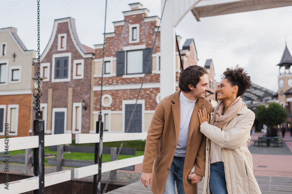 Fototapeta premium joyful multiethnic couple in coats holding hands and smiling near shopping center.