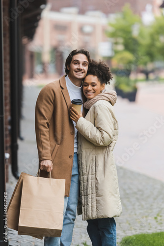 happy african american woman holding paper cup near smiling boyfriend with shopping bags. © LIGHTFIELD STUDIOS