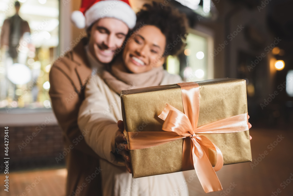 blurred and happy african american woman holding gift box near boyfriend in santa hat. blurred and happy african american woman holding gift box near boyfriend in santa hat.