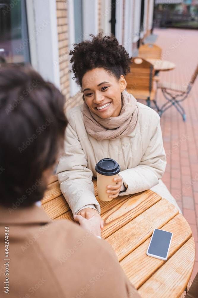 happy african american woman holding hands with blurred boyfriend near smartphone and paper cup. happy african american woman holding hands with blurred boyfriend near smartphone and paper cup.