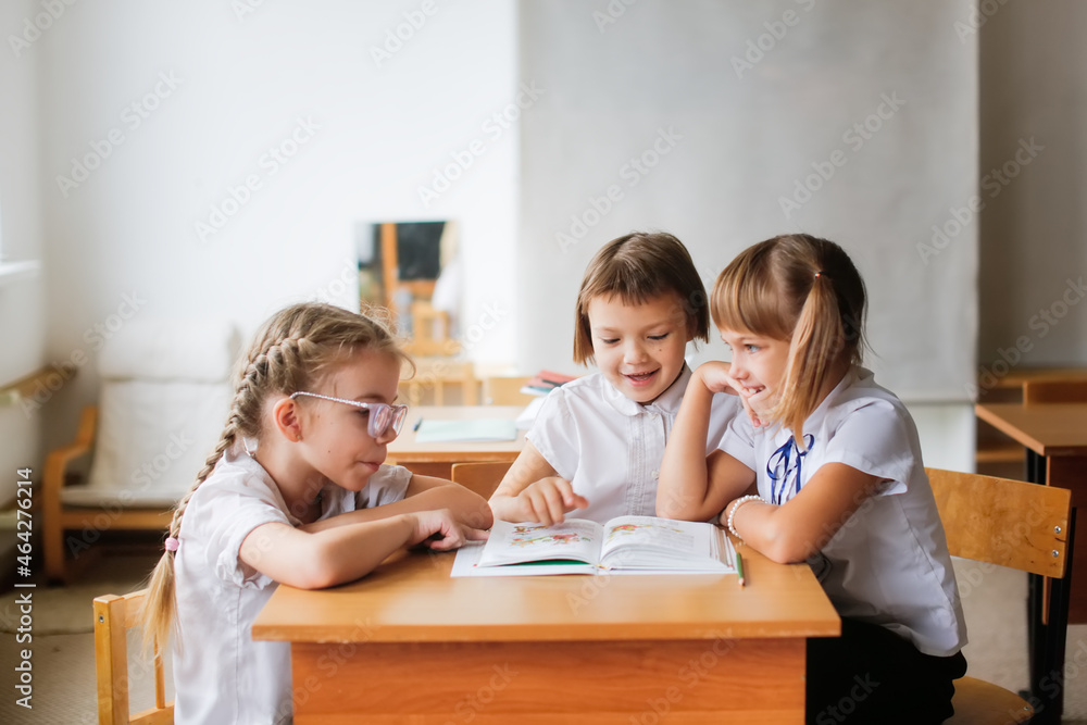 Three girlfriends laughing, classmates read a book together, solve problems. Mutual assistance at school, children sit studying at desks, school learning concept Three girlfriends laughing, classmates read a book together, solve problems. Mutual assistance at school, children sit studying at desks, school learning concept