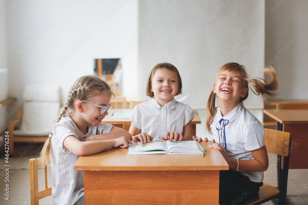 Three girlfriends laughing, classmates read a book together, solve problems. Mutual assistance at school, children sit studying at desks, school learning concept Three girlfriends laughing, classmates read a book together, solve problems. Mutual assistance at school, children sit studying at desks, school learning concept