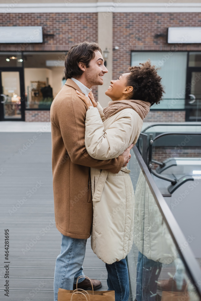 side view of happy man hugging cheerful african american girlfriend in shopping mall.