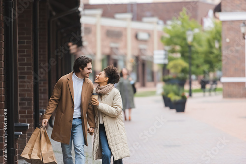 joyful multiethnic couple in coats holding hands while looking at each other and walking with purchases. © LIGHTFIELD STUDIOS