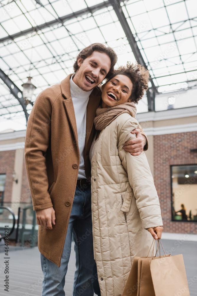 joyful man hugging cheerful african american girlfriend with shopping bags in mall.