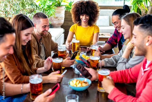 young multiethnic group of friends holding smartphone on social media having fun together drinking happy hour beer at bar restaurant sitting on table indoor. group of diverse people cheering together.