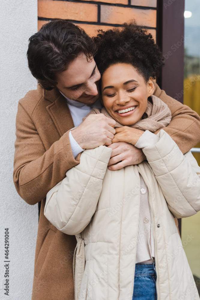 joyful man embracing cheerful african american girlfriend near building outdoors. joyful man embracing cheerful african american girlfriend near building outdoors.