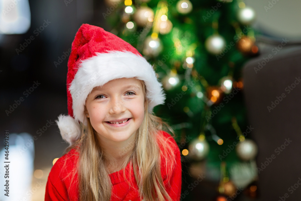 Cute blonde caucasian girl 7 years old sitting on floor near Christmas tree in Santa hat and smiling. Bedroom decorated with garlands. Xmas celebrate, happy new year 2022. Time to make wishes Cute blonde caucasian girl 7 years old sitting on floor near Christmas tree in Santa hat and smiling. Bedroom decorated with garlands. Xmas celebrate, happy new year 2022. Time to make wishes