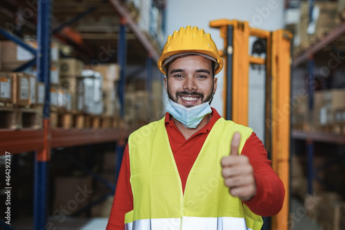 Young hispanic man working inside warehouse while wearing safety face mask under chin and showing thums up on camera photo