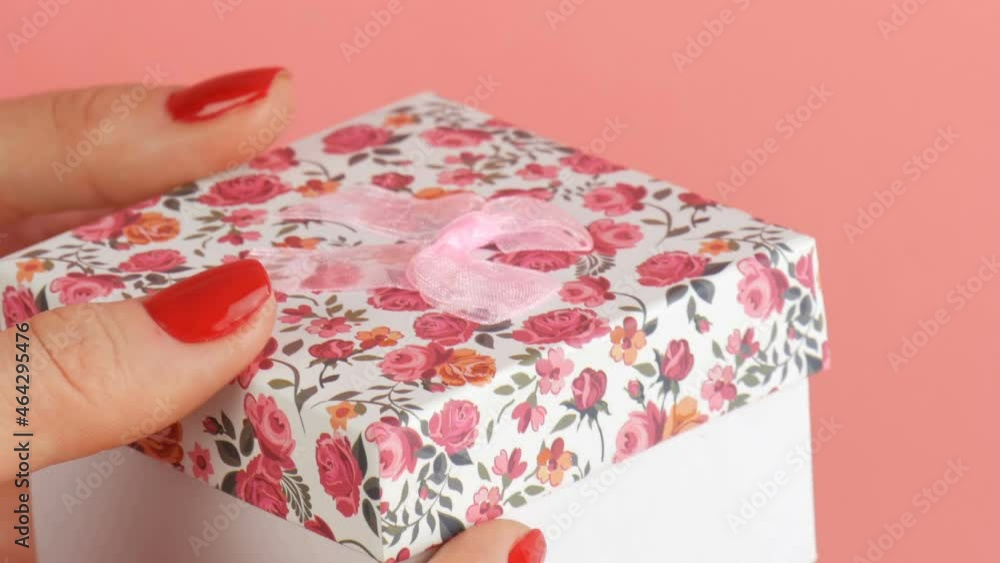 A small gift box with a floral print and a pink bow that is held by the hands of woman whose nails are painted with red nail polish on a pink background. A small gift box with a floral print and a pink bow that is held by the hands of woman whose nails are painted with red nail polish on a pink background.