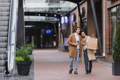 full length of african american woman holding shopping bags and pointing while walking with happy boyfriend in coat in mall.