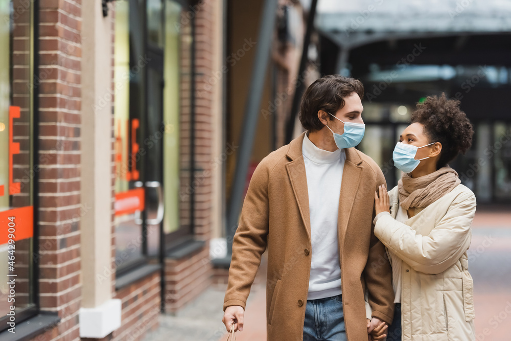 multiethnic couple in medical masks holding hands while walking in shopping mall.