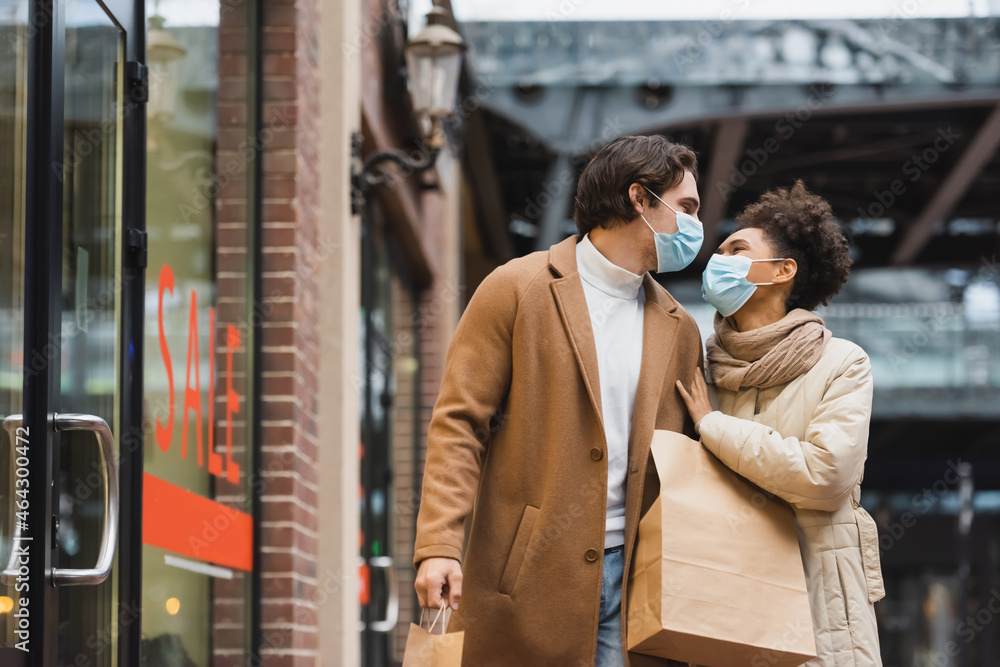 young interracial couple in medical masks looking at each other and walking with shopping bags in mall.