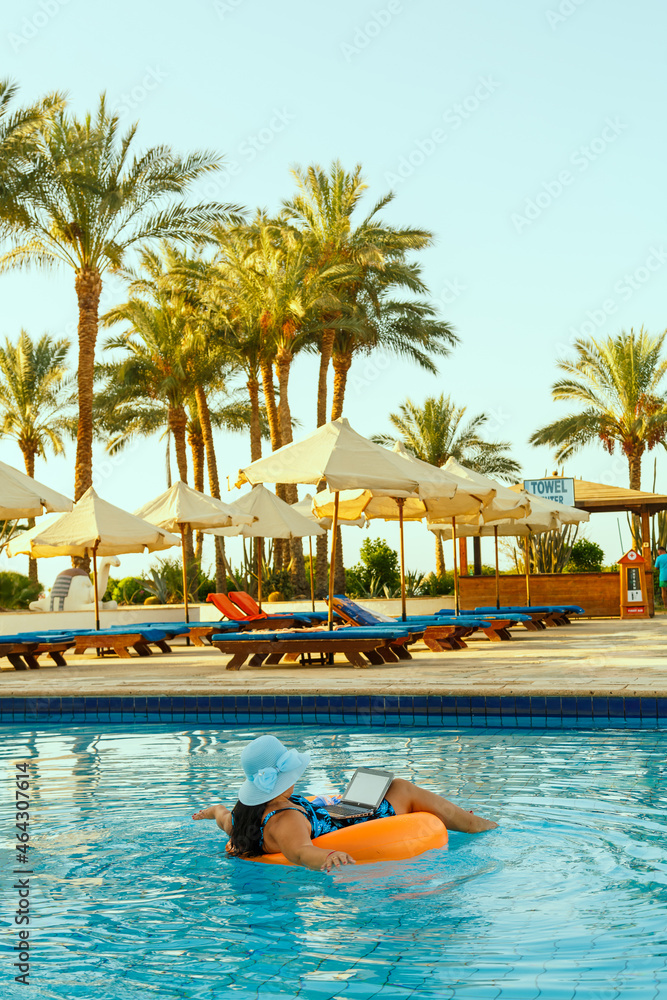 Obraz premium A woman in a sun hat in the pool in a swimming circle with a laptop on the background of palm trees and sun loungers.