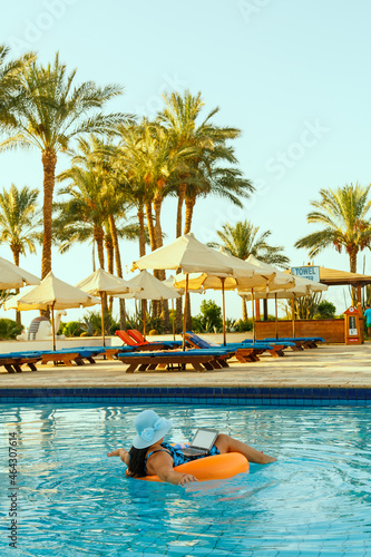 A woman in a sun hat in the pool in a swimming circle with a laptop on the background of palm trees and sun loungers. © finist_4