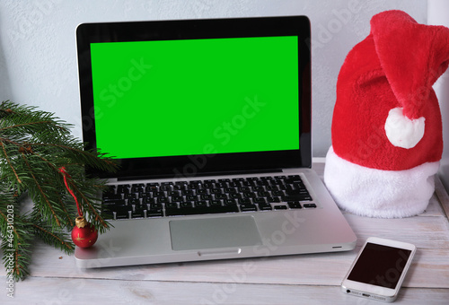 Christmas sales and online shopping for gifts.Laptop with green blank screen, smartphone, Santa hat, fir branches and red ball on wooden background.Selection presents of for new year.