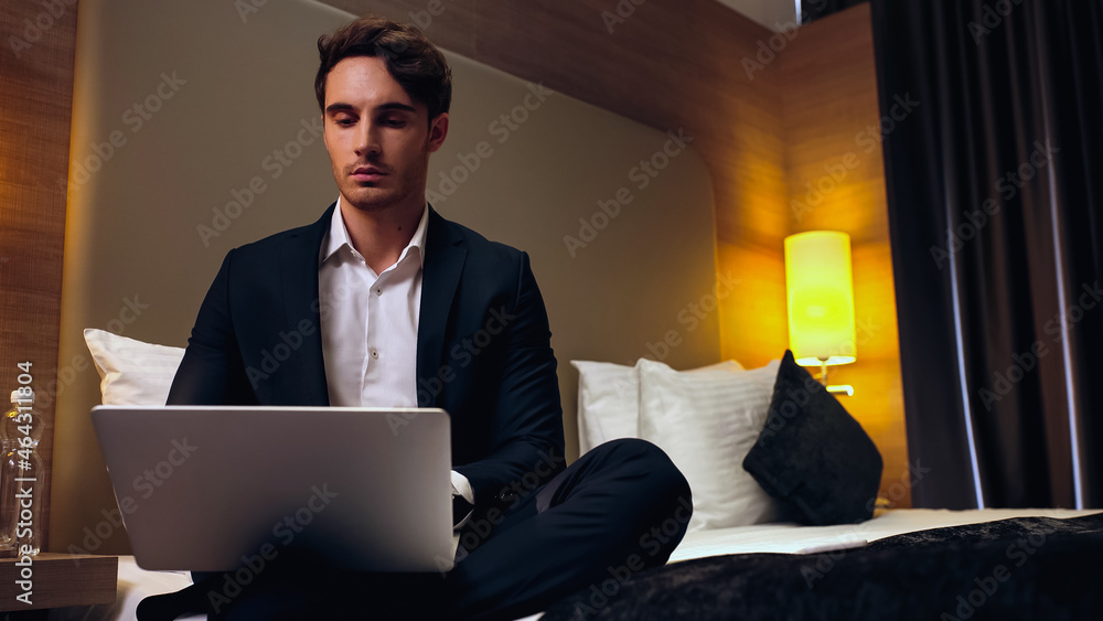 young serious businessman sitting on bed and using laptop in hotel room. young serious businessman sitting on bed and using laptop in hotel room.