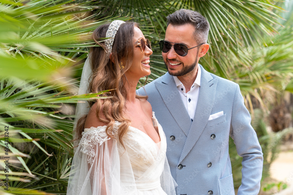 Happy newlywed couple standing near palms in garden Happy newlywed couple standing near palms in garden