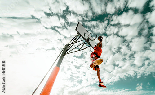 Street basketball player making a powerful slam dunk on the court - Athletic male training outdoor on a cloudy sky background - Sport and competition concept 