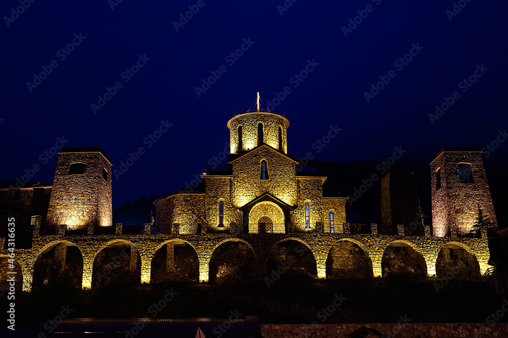 The building of an old stone monastery that looks like a castle. At night, the building is illuminated in bright yellowish colors